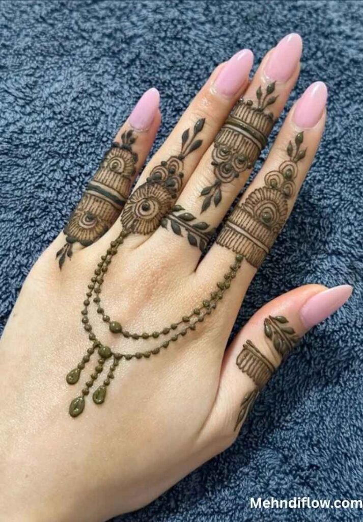 Hand with pink nails featuring ring-style finger mehndi connected by draped dotted chains.
