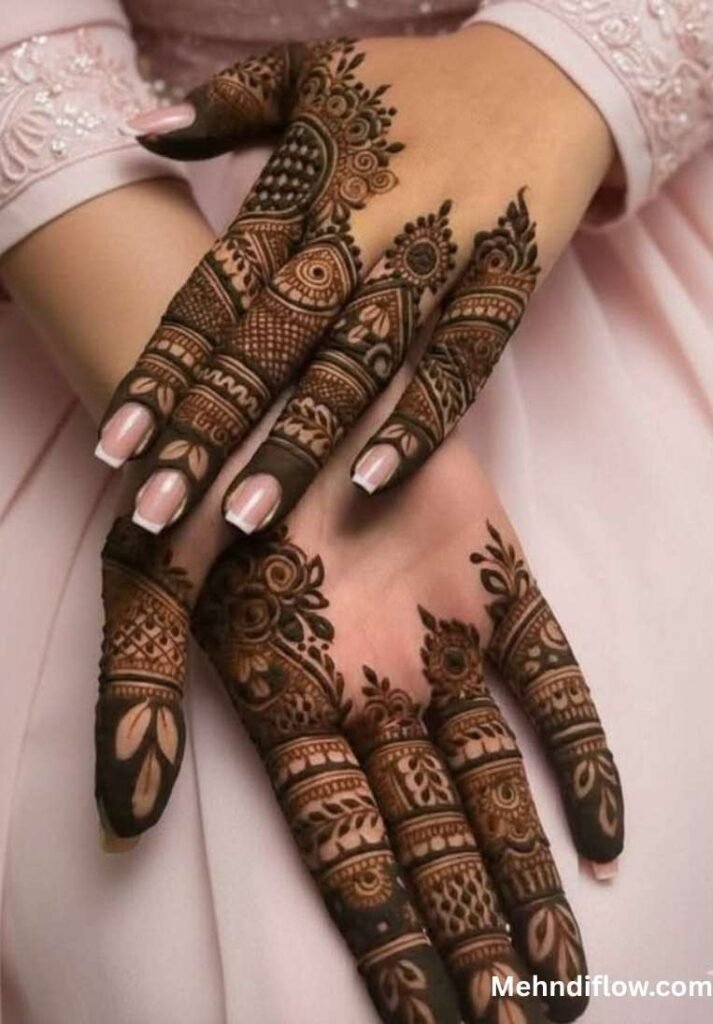 Two hands resting on pink fabric displaying dense, intricate finger mehndi with floral and mesh patterns.