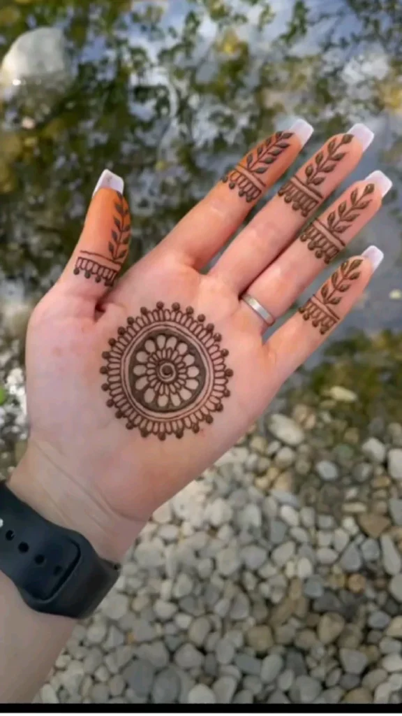 A palm-up hand featuring a central circular mandala mehndi design with a flower in the middle, delicate leafy trails climbing each finger, and a single floral wrist band.