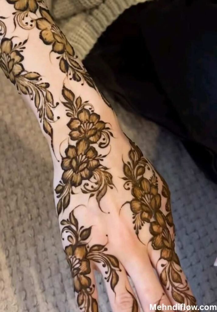 A close-up of a hand showcasing a flowing Arabic fusion mehndi pattern characterized by bold floral motifs and elegant vine-like strokes that extend across the back of the hand.