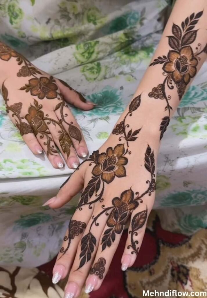 Elegant floral Arabic mehndi design trailing across the back of two hands, featuring dark-shaded flower motifs and prominent leaf patterns extending down the fingers and wrist.
