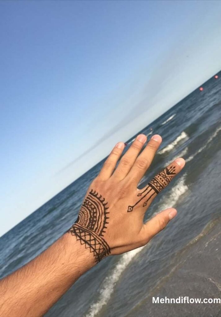 A hand shown against an ocean backdrop, featuring a modern, linear Arabic-style mehndi design with a structured wrist cuff and a minimalist pattern extending onto the index finger.