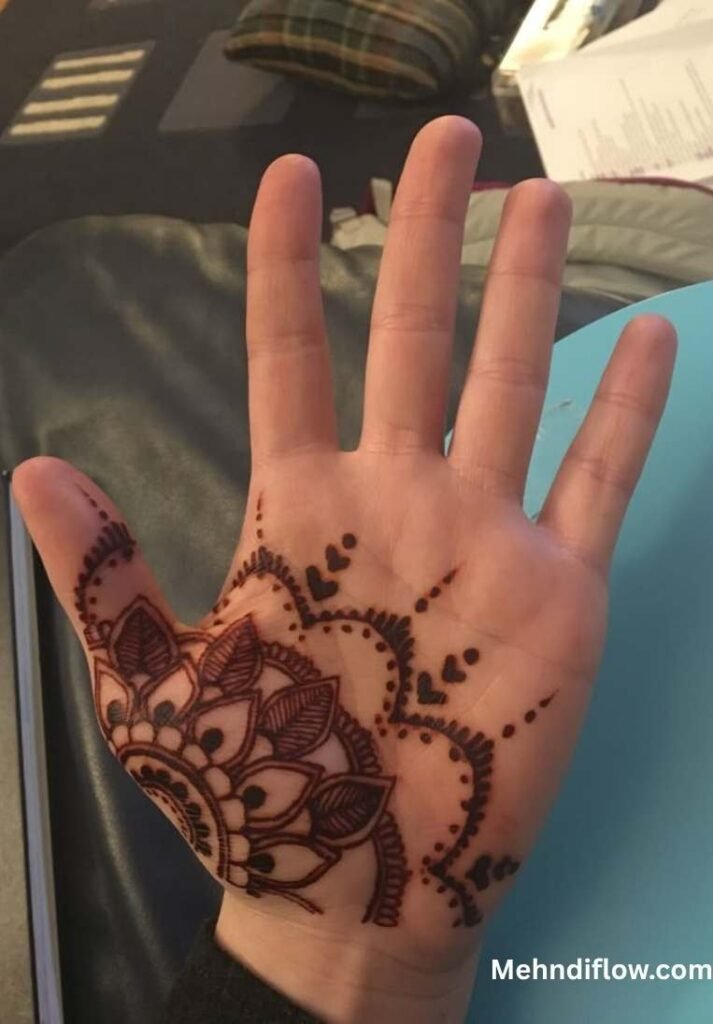 A palm displaying a traditional Arabic-style mandala mehndi design, featuring bold floral petals, scalloped edges, and small heart accents.