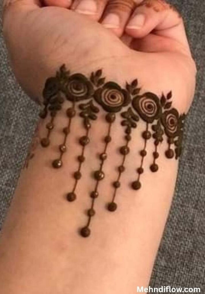 Close up of a wrist featuring a bold bracelet-style mehndi design composed of a neat row of floral swirls and tiny leaves, with delicate hanging beaded jhumka trails extending down the forearm.