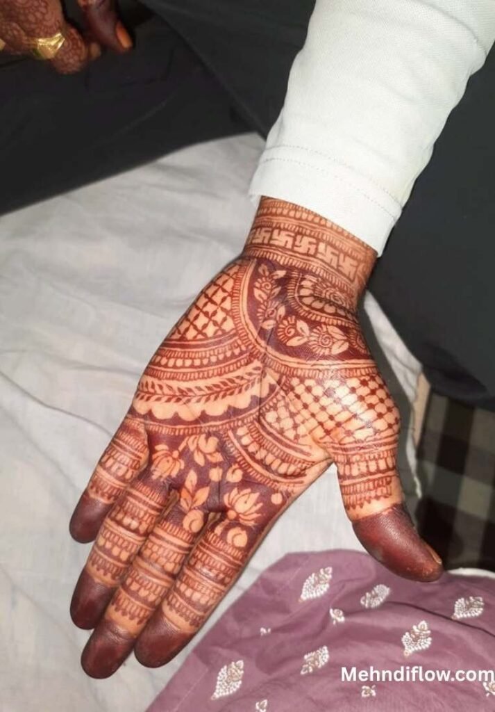A hand featuring an elaborate, full-coverage groom's mehndi design with dense traditional patterns, including a prominent wrist band with geometric swastika motifs.