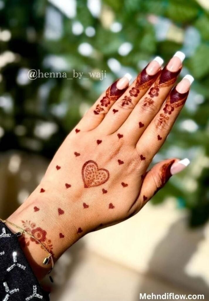 Close up of the back of a hand featuring a central heart-shaped frame filled with detailed intricate henna mesh patterns, surrounded by a minimalist scattering of tiny heart motifs, detailed floral caps near the fingernails, and a matching wrist band design