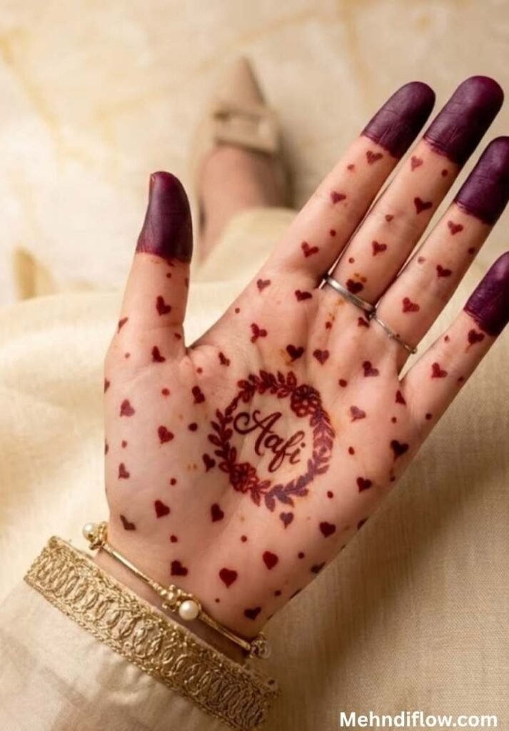 Close up of an open palm featuring a central circular floral wreath enclosing the name 'Aafi' in cursive script, surrounded by a minimalist pattern of tiny scattered heart shapes across the entire palm and fingers with darkly stained fingertips