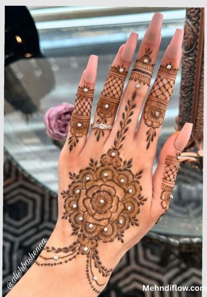 A close-up view of a hand displaying a jewelry-inspired mehndi design, highlighting a central floral motif combined with net patterns on the fingers, accented by small, jewel-like decorative dots.