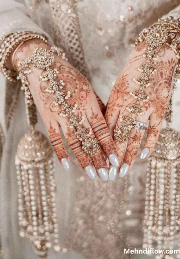 A pair of hands adorned with intricate bridal mehndi that mimics the appearance of jewelry, featuring elaborate patterns complemented by decorative hanging hand accessories.