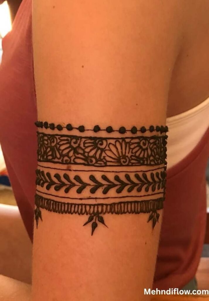 Geometric bracelet style arm mehndi design wrapped around the upper bicep, featuring horizontal bands of detailed floral patterns, leafy vines, and delicate hanging leaf accents.
