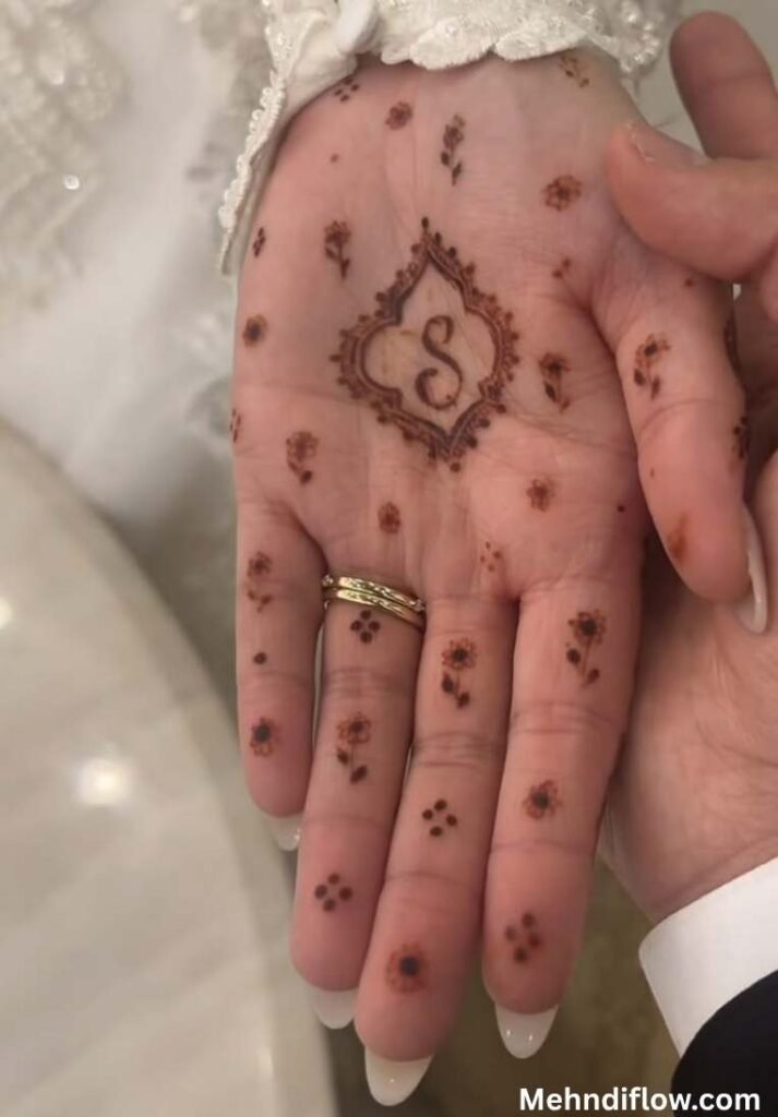 Close up of a person's palm with a detailed diamond-shaped frame enclosing the letter 'S' in cursive script, surrounded by a minimalist pattern of tiny individual flower buds scattered across the palm and fingers.