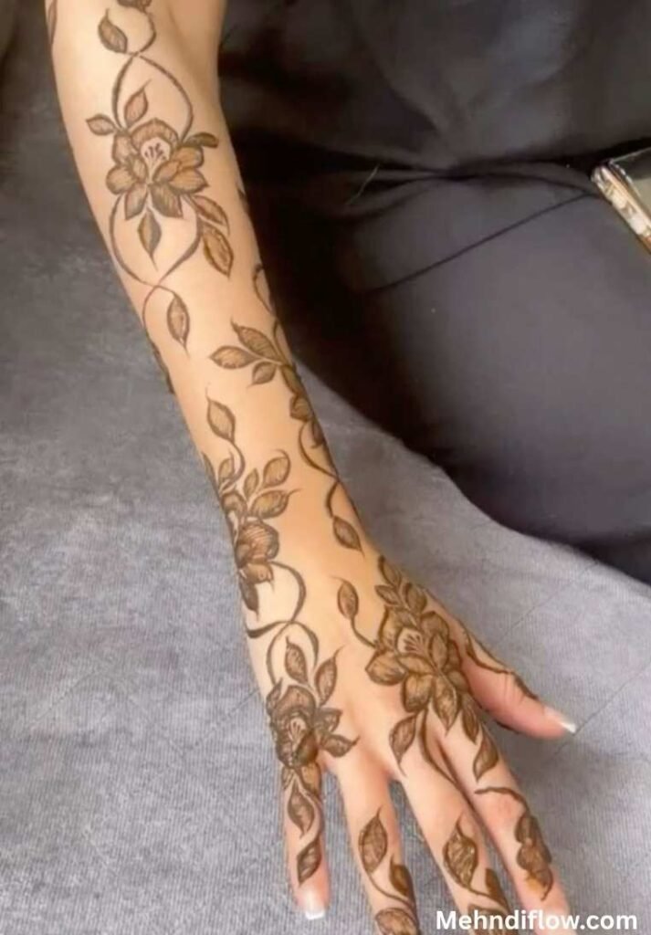 A forearm and hand adorned with flowing tropical leaf and floral henna patterns.