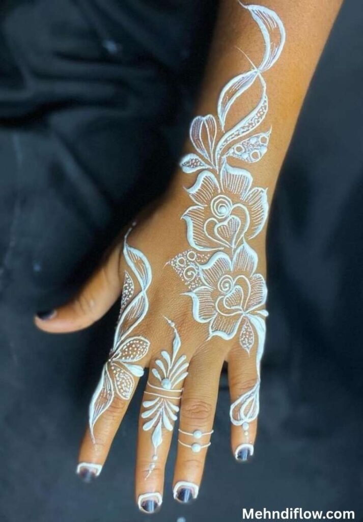 Bold floral white mehndi design featuring large shaded roses, flowing leafy vines, and delicate finger accents extending from the wrist down the back of the hand.