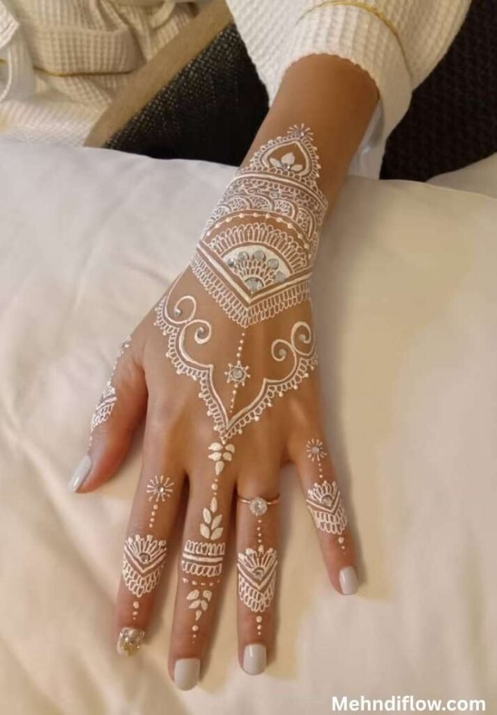 Jewelry-style white and gold henna design resembling a traditional hand harness, showcasing a geometric central motif, dotted chain links, and intricate patterned finger cuffs.